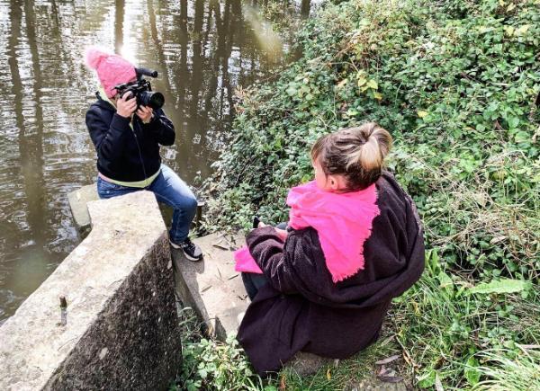 Two women sitting by a river, one of them is being filmed and the other one is holding a camera