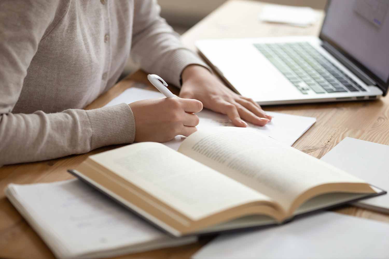 Someone making notes from a book with a laptop