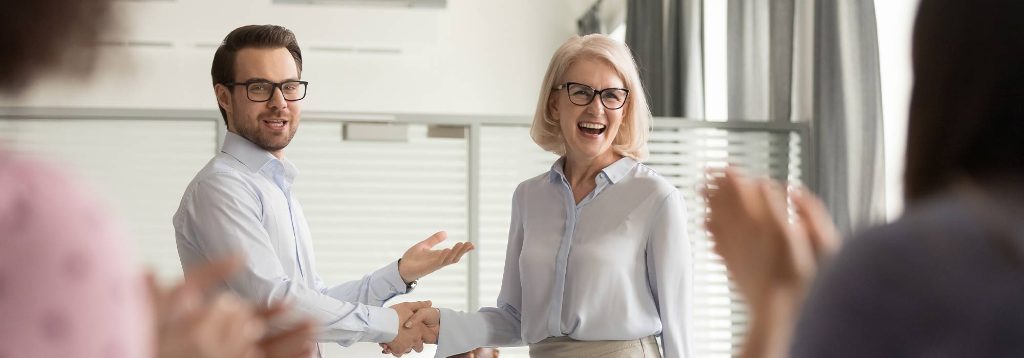 Two people in white shirts and business attire shaking hands and laughing in an office filled with people