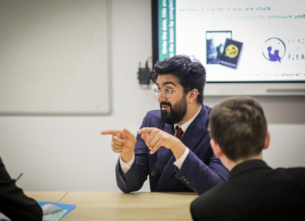 Person with dark hair and beard at front of classroom in suit