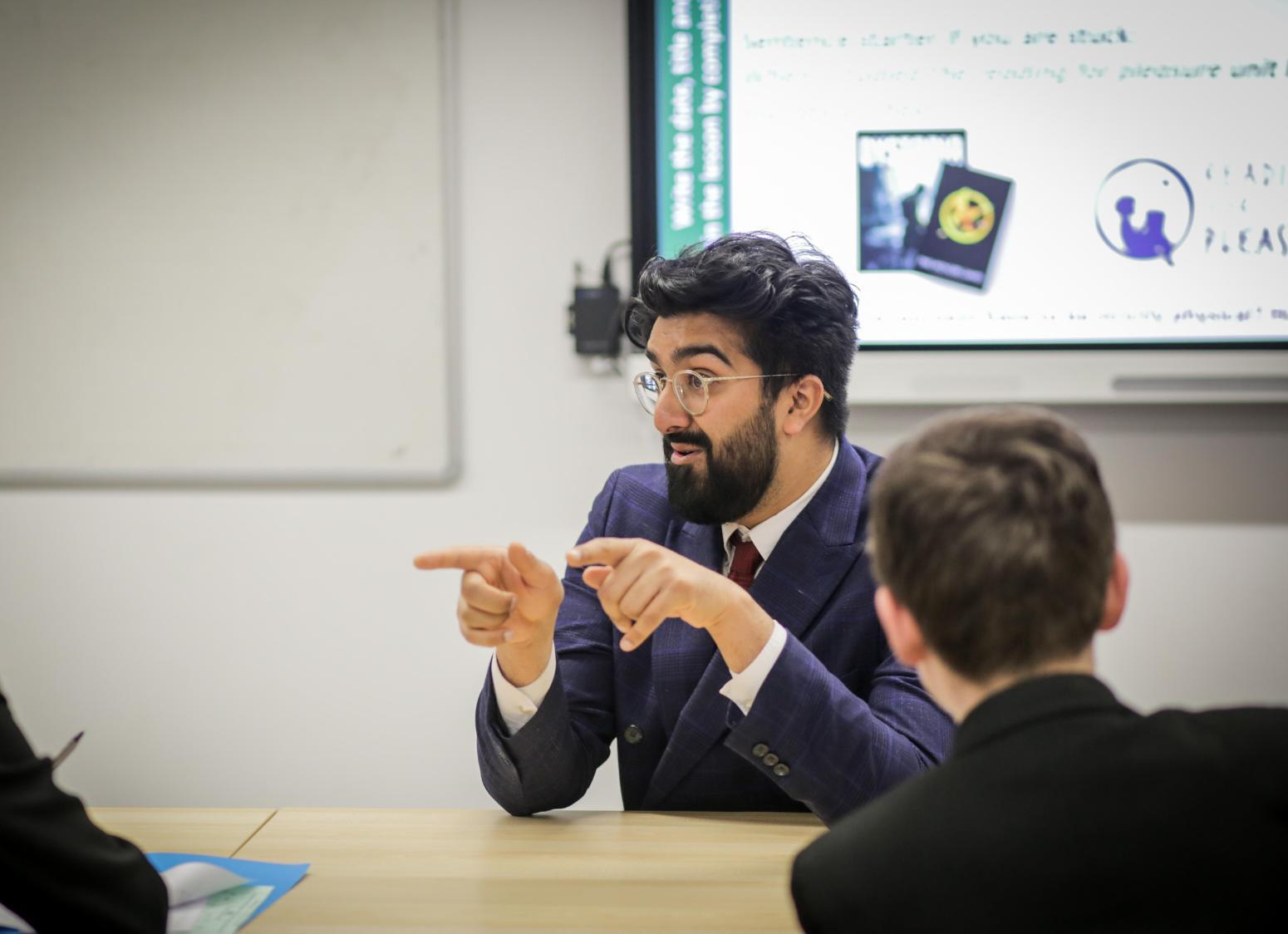 Person with dark hair and beard at front of classroom in suit