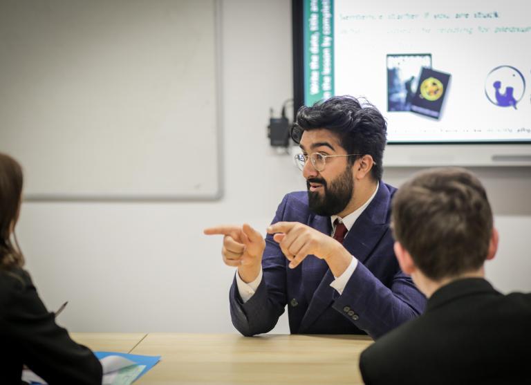 Person with dark hair and beard at front of classroom in suit