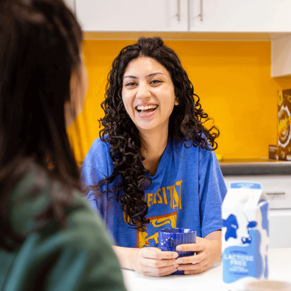 A student sits in an accommodation kitchen, laughing with a cup of tea.