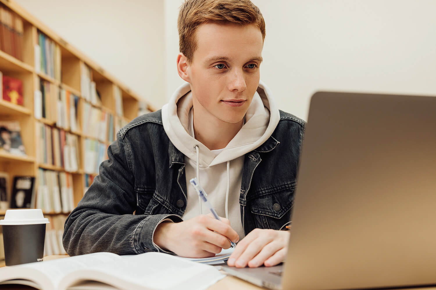 A male-presenting student sat in a library, writing in a notebook whilst looking at his laptop.