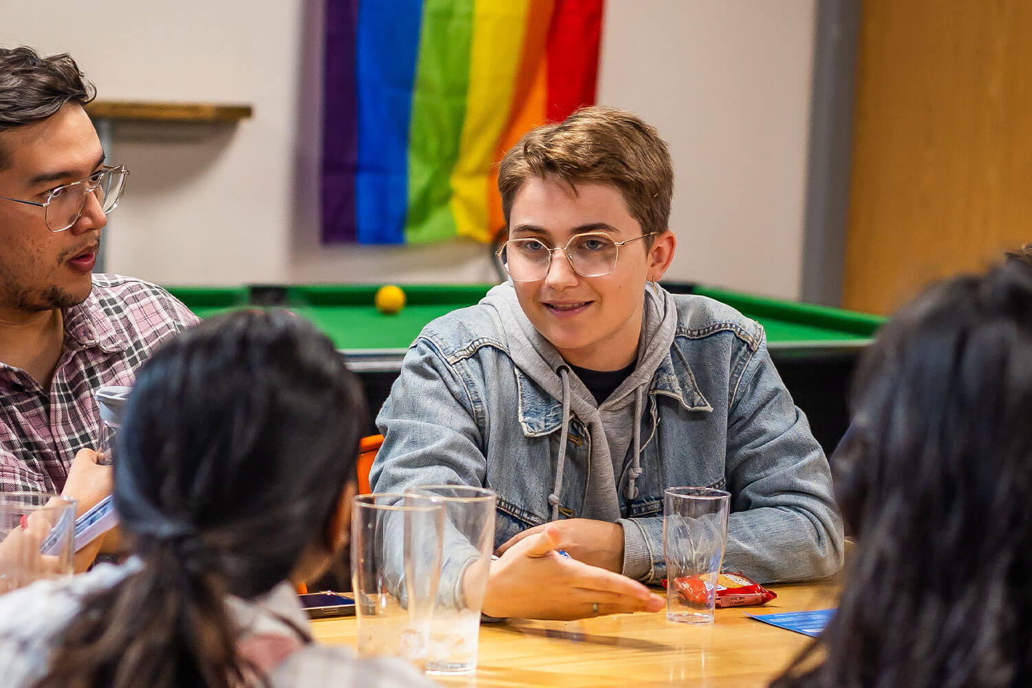 Four students sat together in the Student Union with a pool table and pride flag in the background