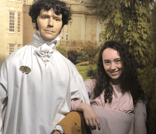 A student stands smiling in regency dress linking arms with a mannequin dressed in regency clothing