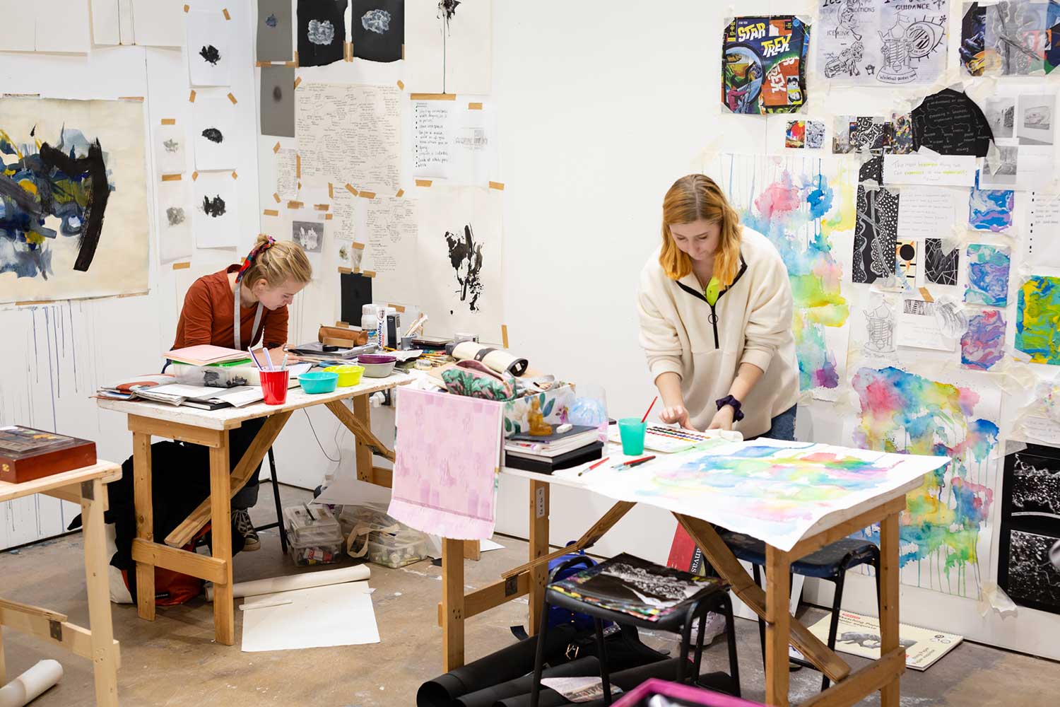 Two students stand at their desks, preparing pieces of artwork