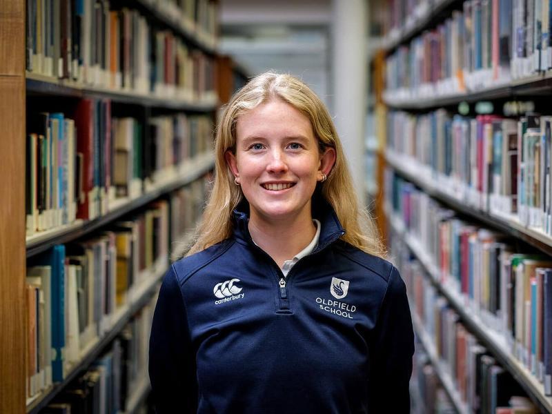 A young woman wearing school-branded PE kit smiling at the camera with a library in the background