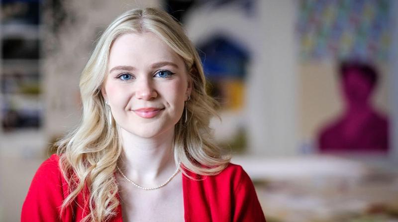 Woman with blue eyes, blonde curls and a bright red blazer sitting in a studio environment