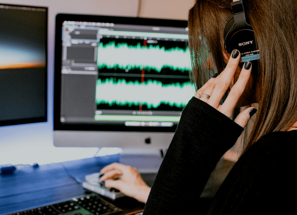 a woman sits at her desk with headphones on. One of her two computer monitors shows soundwaves from the music she is listening to.