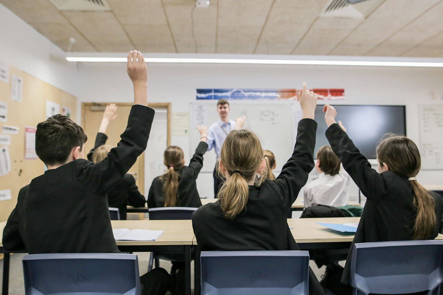 Teacher at the front of a class in which many students have their hands raised