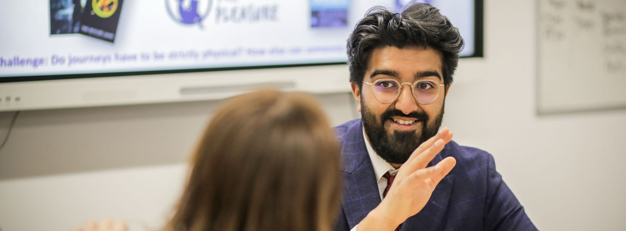 Smiling trainee teacher with a dark blue suit gestures while talking to a school pupil