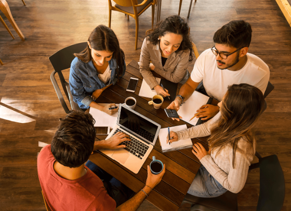 Five students sit around a table drinking coffee and writing in notebooks