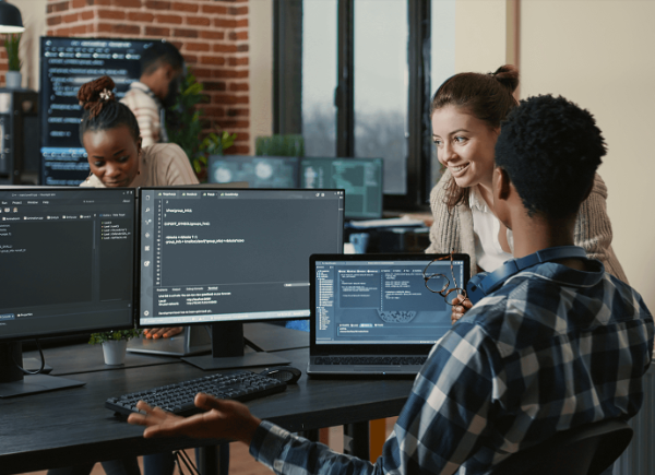 Two coworkers look at three computer screens of coding language and discuss