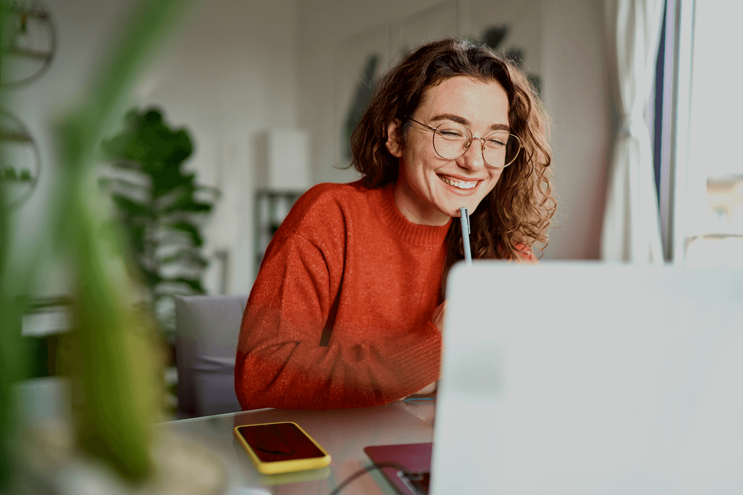 Happy young woman using laptop sitting at desk writing notes whilst looking at laptop screen learning online.