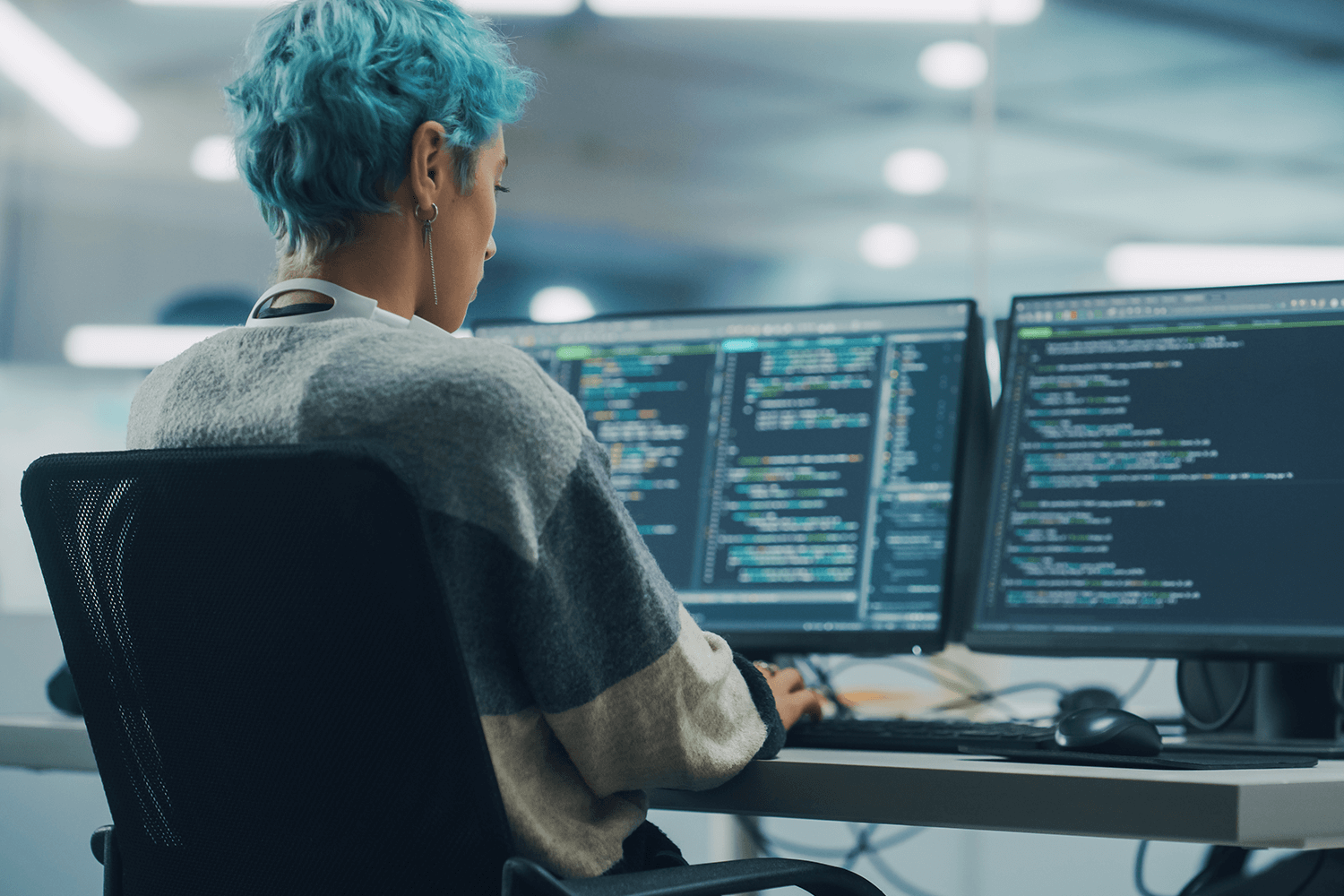 A worker sits at their desk coding on two monitors.