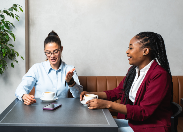 Two coworkers sit next to each other at a cafe. They are talking to each other and drinking cups of coffee.