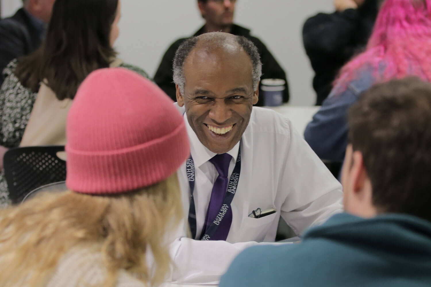 Smiling man in smart clothing chats to a woman in a pink hat and a man in a blue top at a teacher education evening event