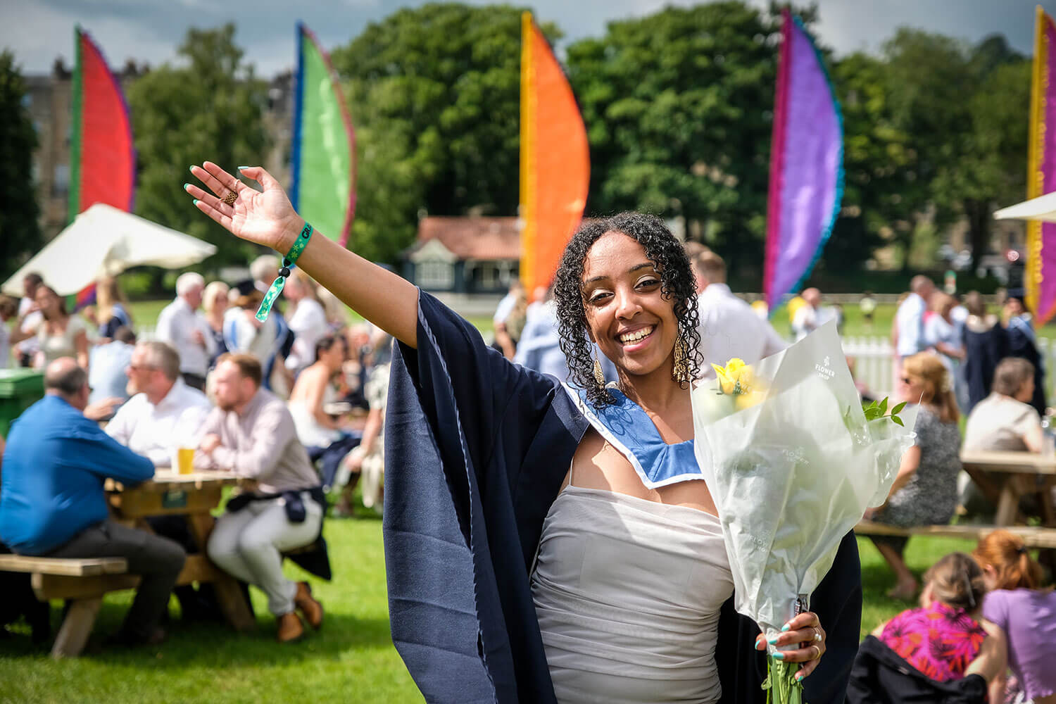 Graduate holding a bunch of flowers and holding her arm out in celebration at an outdoor event with colourful flags in the background