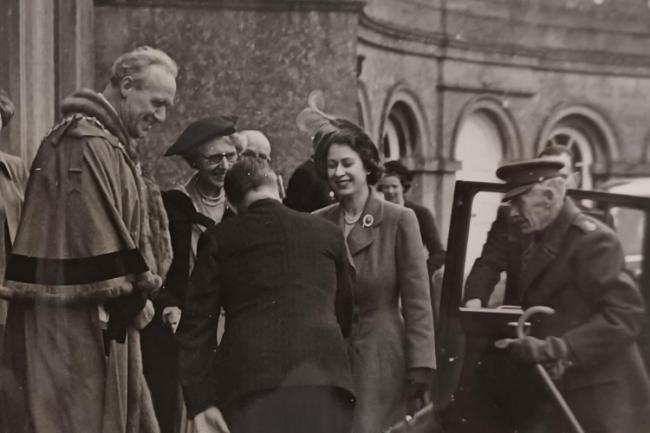 Princess Elizabeth steps out of a car and shakes hands with someone bowing, people gather around her. The walls of Main House at Newton Park can be seen in the background.