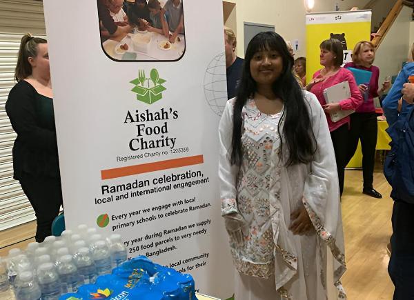 Student Aishah Ahmed stands next to a banner that says Aisha's Food Charity