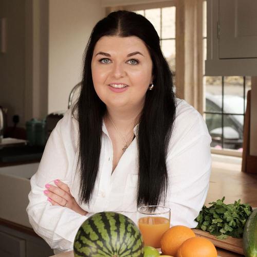 A woman stood in a kitchen posing for photo with a watermelon and two oranges sat on the kitchen top in front of her