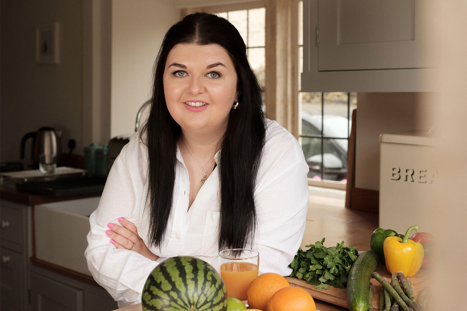 A woman stood in a kitchen posing for photo with a watermelon and two oranges sat on the kitchen top in front of her