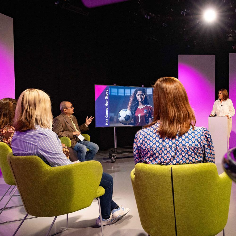 three women and one man sit in a studio like room talking to a woman at a podium, a screen is behind them showing her game her story. 