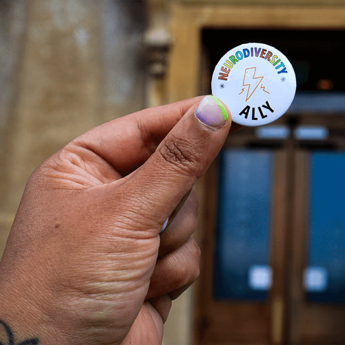 A hand holding up a pin badge. The text on it says Neurodiversity Ally in rainbow letters.