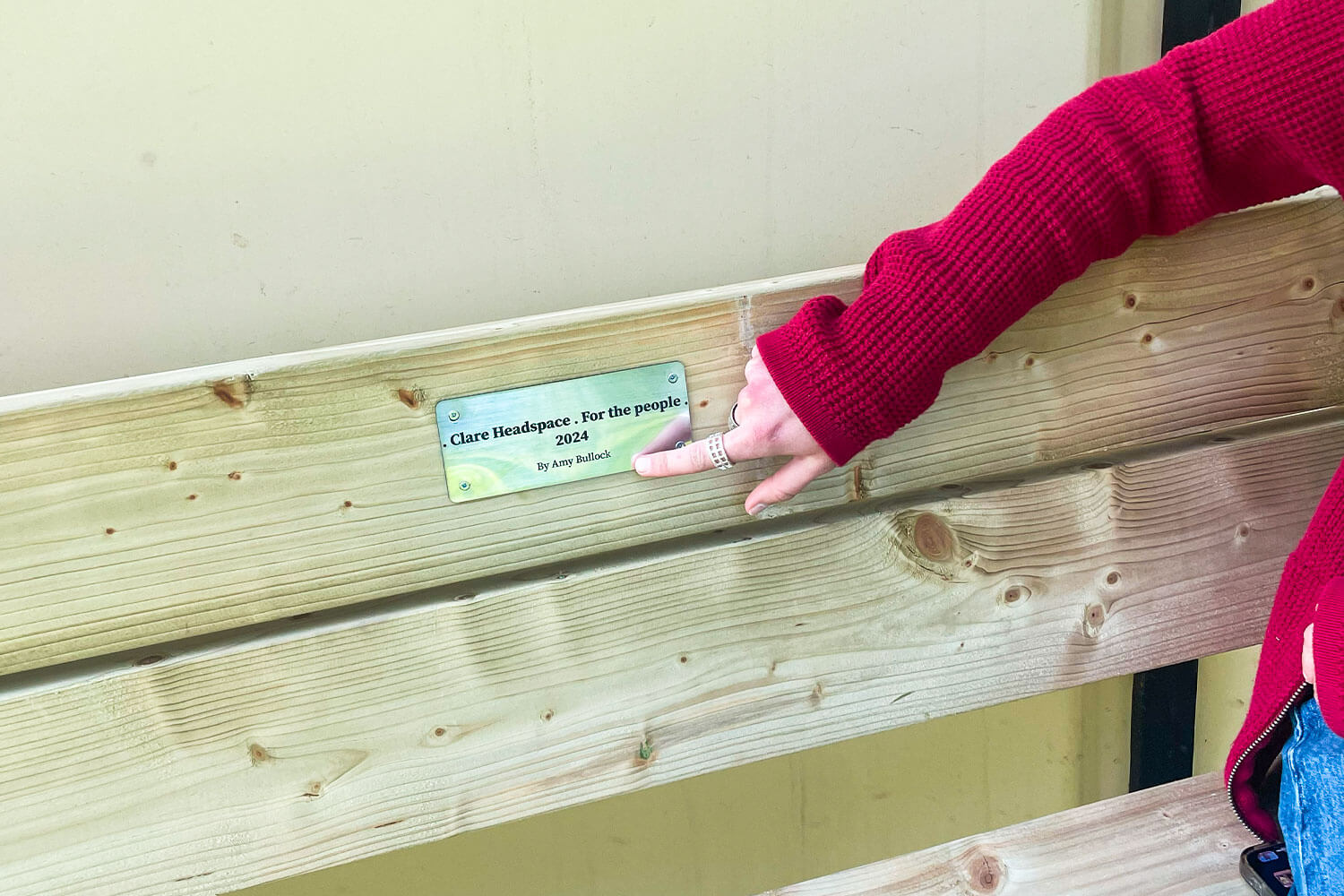 A wooden bench with a finger pointing at a plaque in the centre
