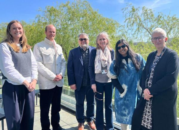 A group of people stand together smiling on balcony. Blue sky and trees are in the background.
