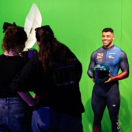 A bobsleigh athlete stood in front of a green screen, posing for two people stood behind a camera