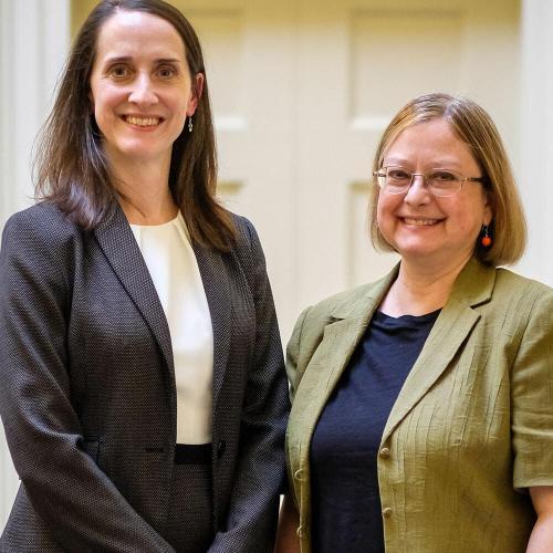 Two women in smart clothing stand together smiling.