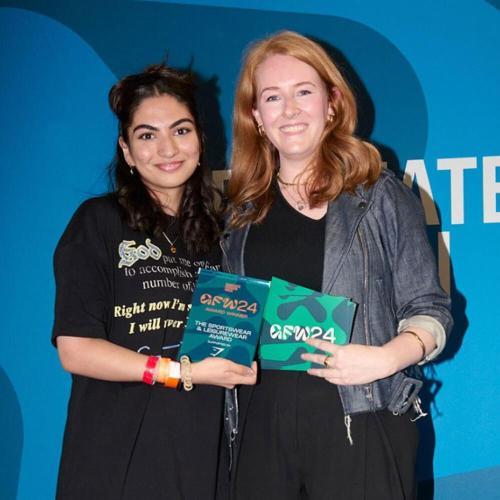 Two people stand together, both holding graduate fashion week awards and smiling at the camera.