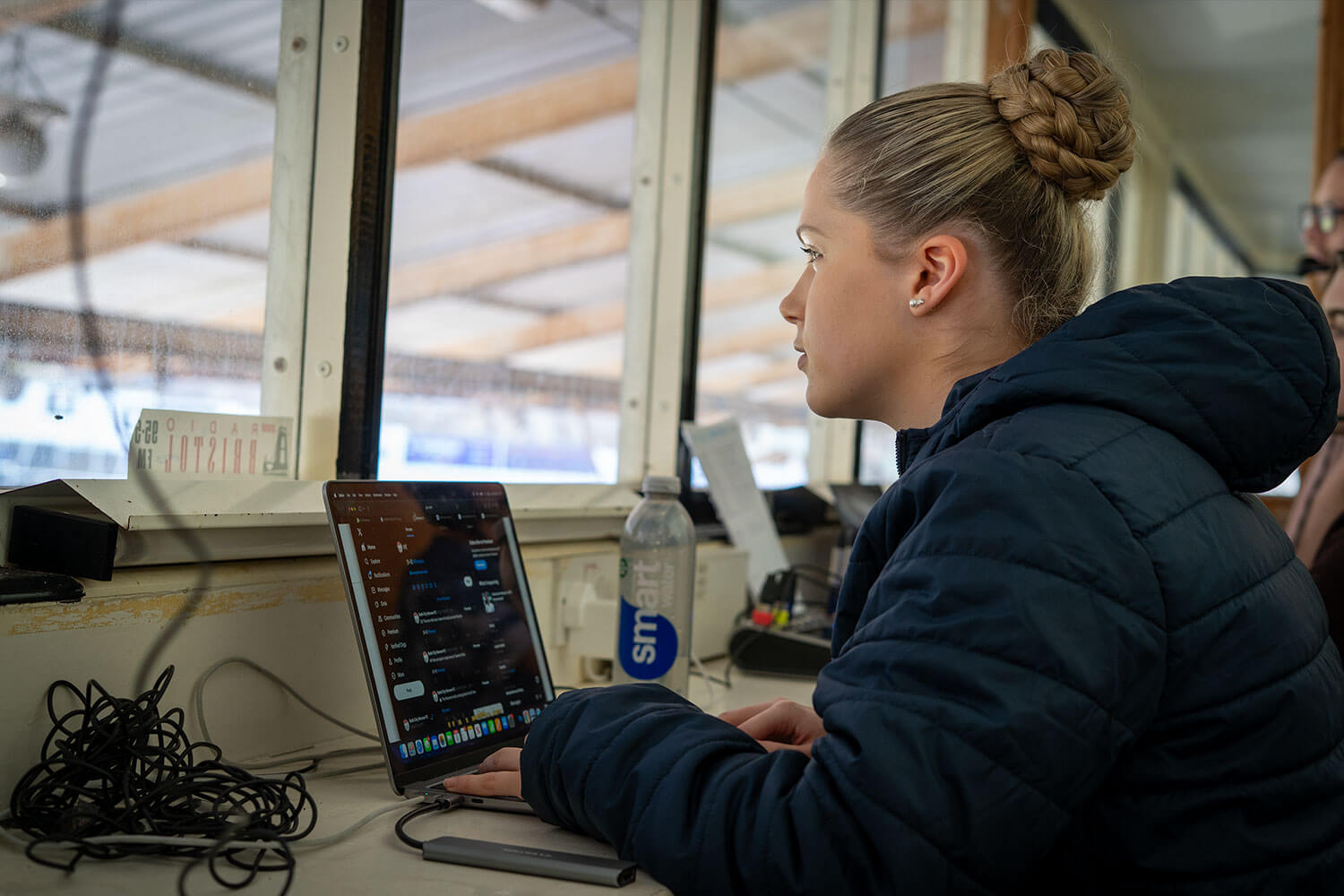 A girl typing on a laptop inside a press box in a football stadium