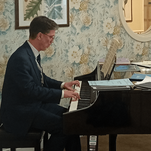 A male student wearing a black suit sits at a piano in a care home playing a piece for the elderly residents.