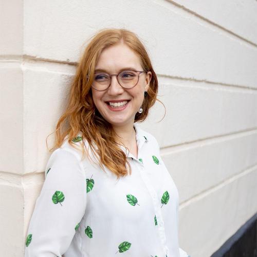 a woman leans on a plain wall, smiling, wearing wearing glasses and a blouse with a leaf pattern.