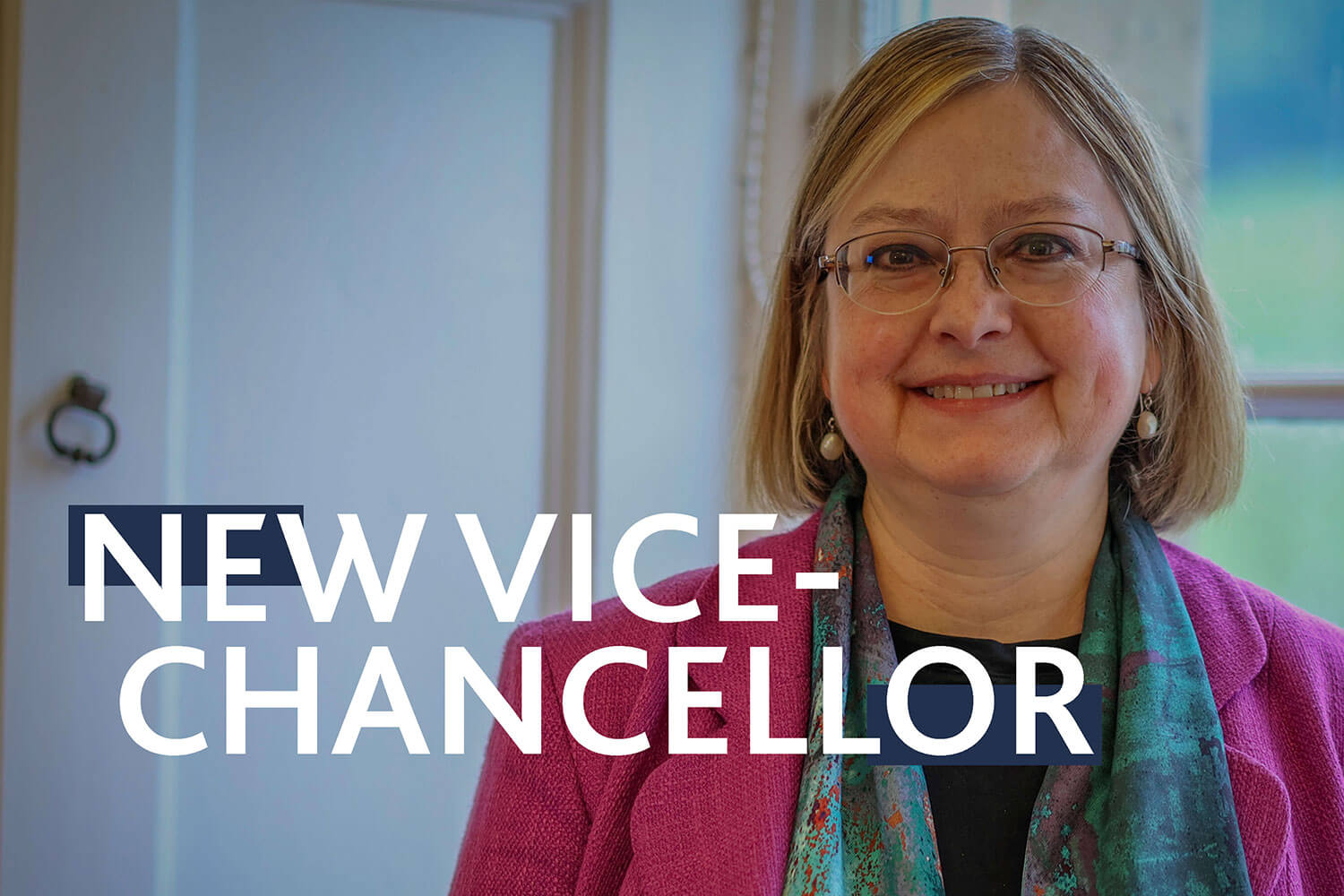 Headshot of Professor Georgina Andrews - woman smiles wearing colourful smart clothing, standing in front of a window with green fields beyond it. Overlay text: New Vice-Chancellor.