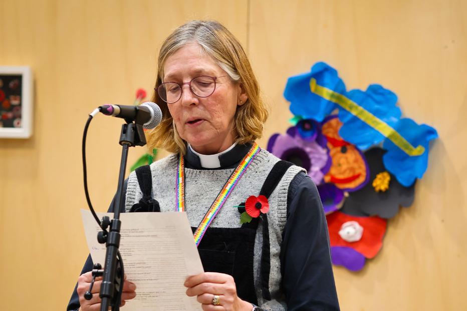 University Chaplain, Katy Garner, leads a service on Remembrance day. She reads form a piece of paper at a microphone, standing in front of a wall of artwork inspired by the remembrance poppy.