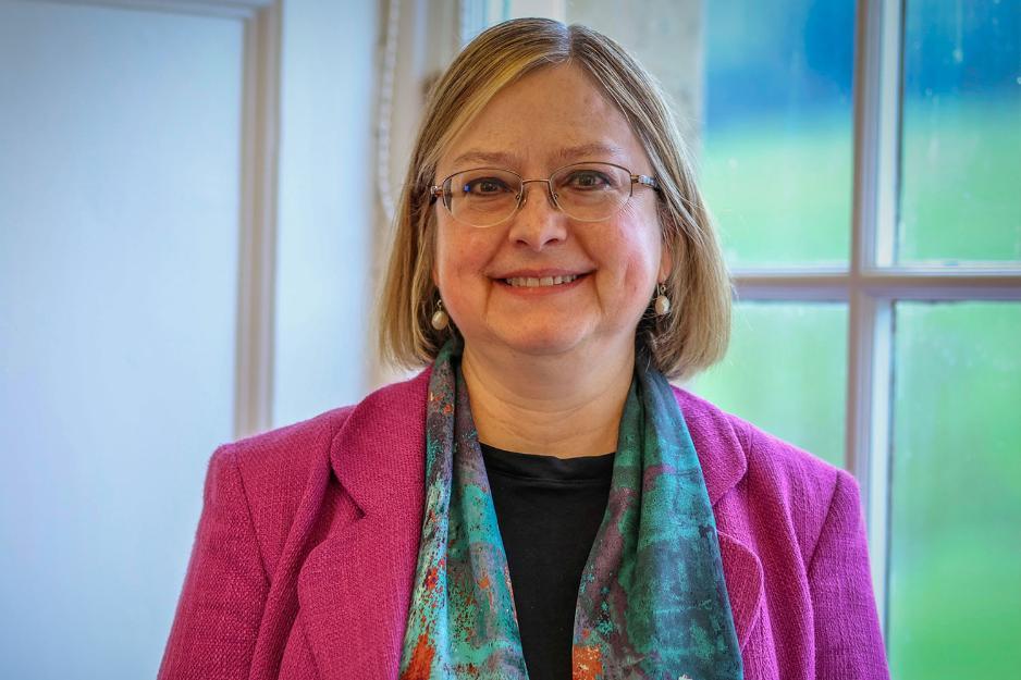 Headshot of Professor Georgina Andrews - woman smiles wearing colourful smart clothing, standing in front of a window with green fields beyond it.