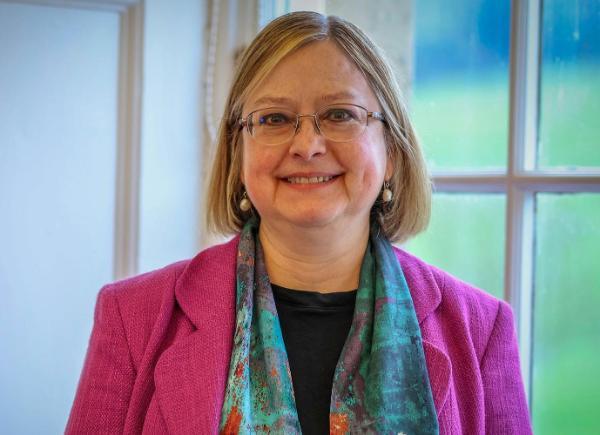 Headshot of Professor Georgina Andrews - woman smiles wearing colourful smart clothing, standing in front of a window with green fields beyond it.