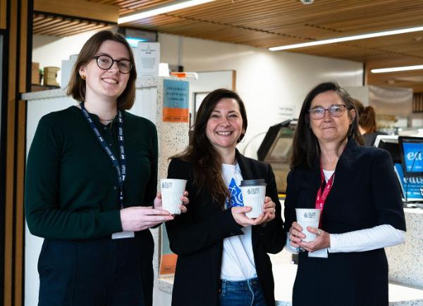 three women smile holding their reusable cups in front of a cafe counter