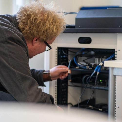 technician crouched down in front of audio visual equipment, looking at wiring.