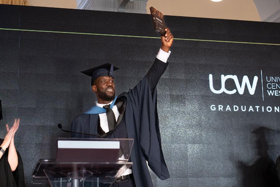 student in graduation cap and gown holds an award up in the air, standing behind a podium on a stage.