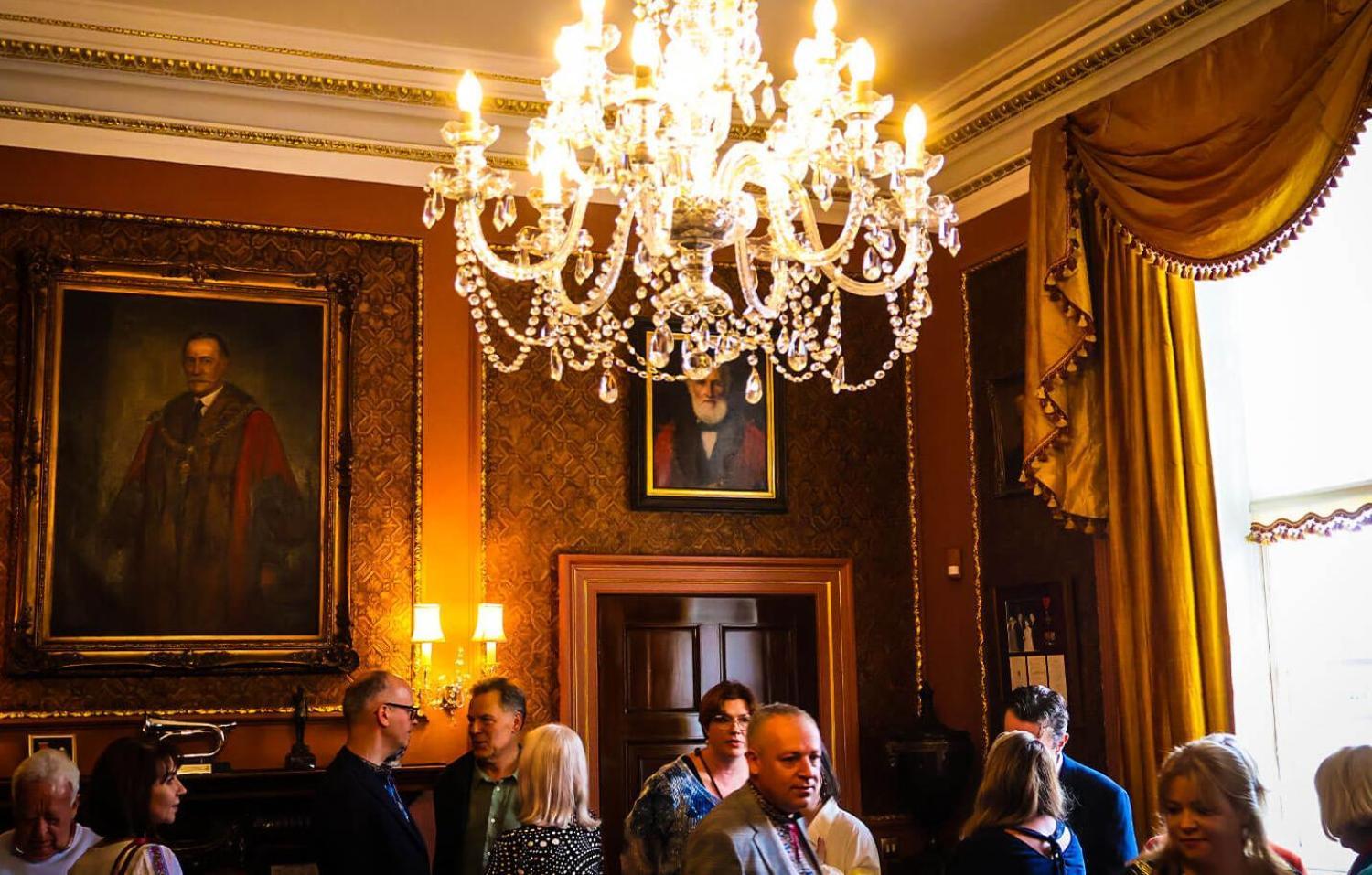 A grand room with paintings on the wall filled with people, with a large chandelier over the top of their heads