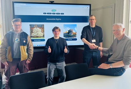 Two students stand smiling in front of a large screen showcasing a website that contains images of ‘Accessible Sights’ in the city of Wells. To the right, Dr Jake Hobbs and a representative from the Accessible Wells group shake hands.