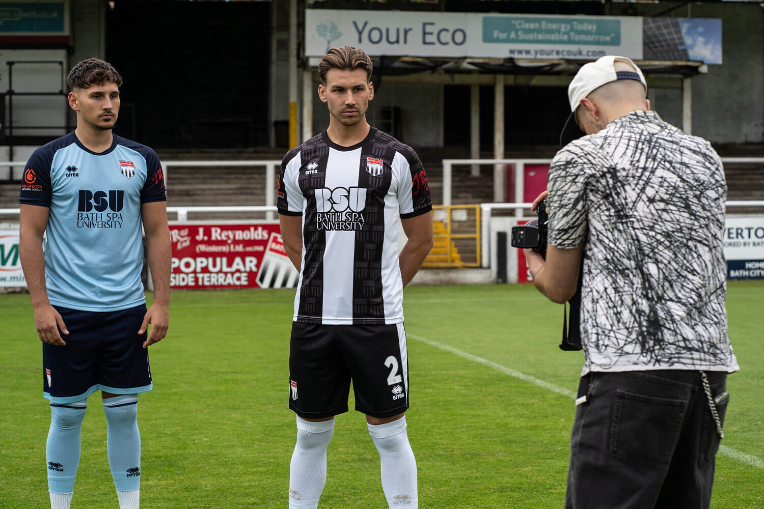 Two football players wearing Bath City shirts, one black and white, one blue, being photographed by a student at Twerton Park football stadium