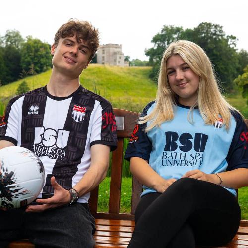 Two students sat on a bench wearing Bath City's new shirts, one black and white and one two-toned navy blue. The pair sit on the bench in front of a green landscape and the male students holds a football