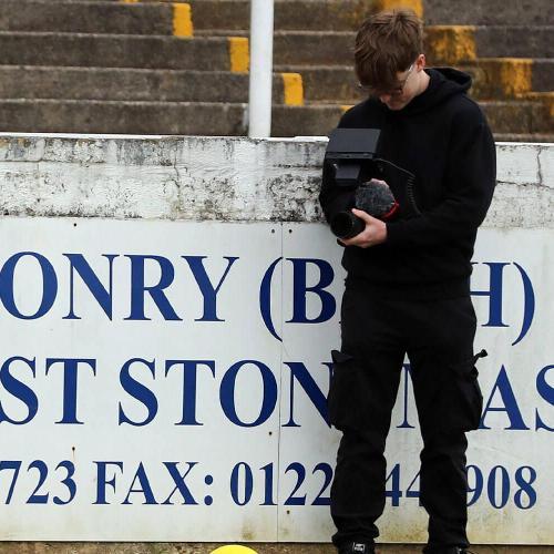 A student holding a camera on a football pitch, stood against a white advertising hoarding at the side of the pitch