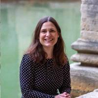 Person with long dark hair sitting in front of Roman baths in a black polka dot dress